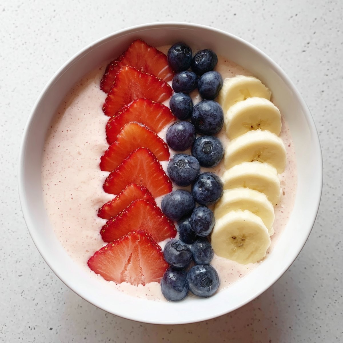 Thick and creamy fruit smoothie bowl topped with sliced strawberries, fresh blueberries, and banana on a light countertop